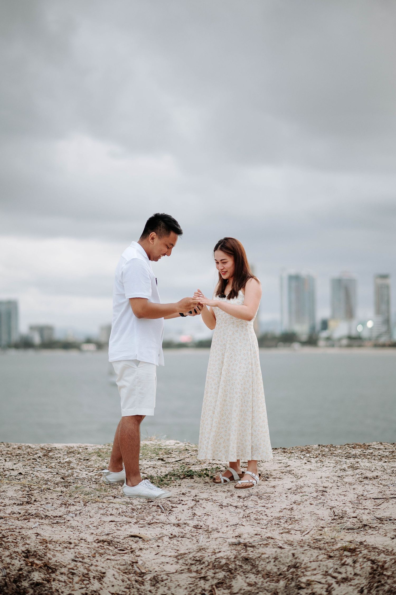 Happy couple examining engagement ring on beach with city view.