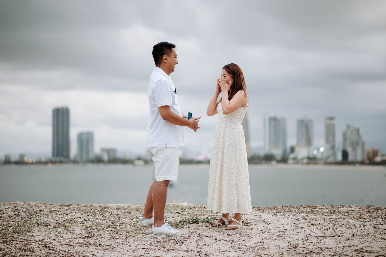 Emotional proposal moment with couple in white outfits by the water.