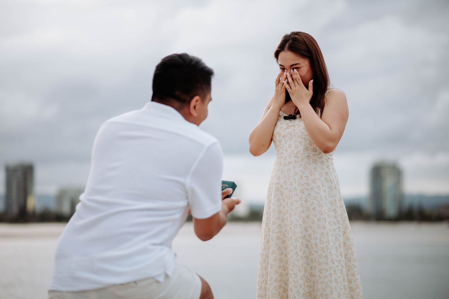 A proposal moment captured on the beach with city skyline in the background.