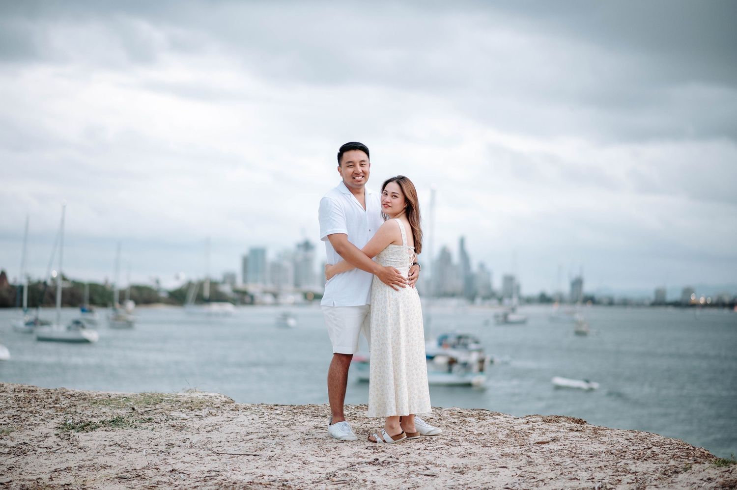 A couple in white outfits poses together on a rocky beach with boats and city skyline in the distance.