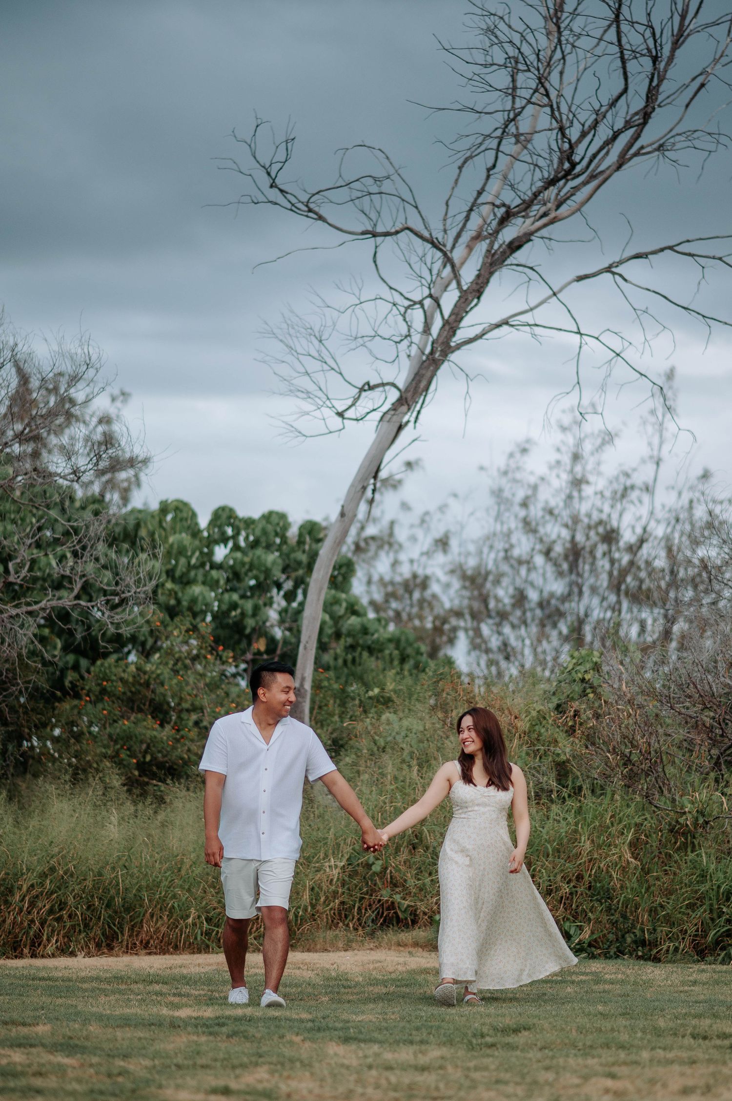A couple holds hands while walking through a grassy area with a bare tree against moody skies.