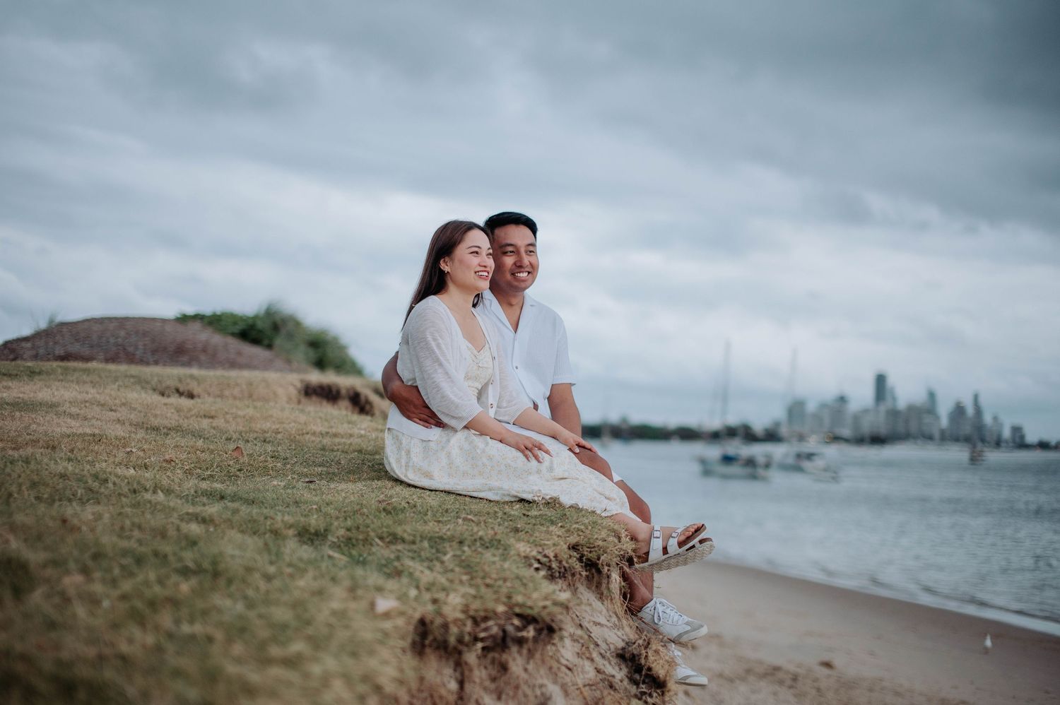 A couple sits together on a coastal cliff overlooking the harbor and city skyline.