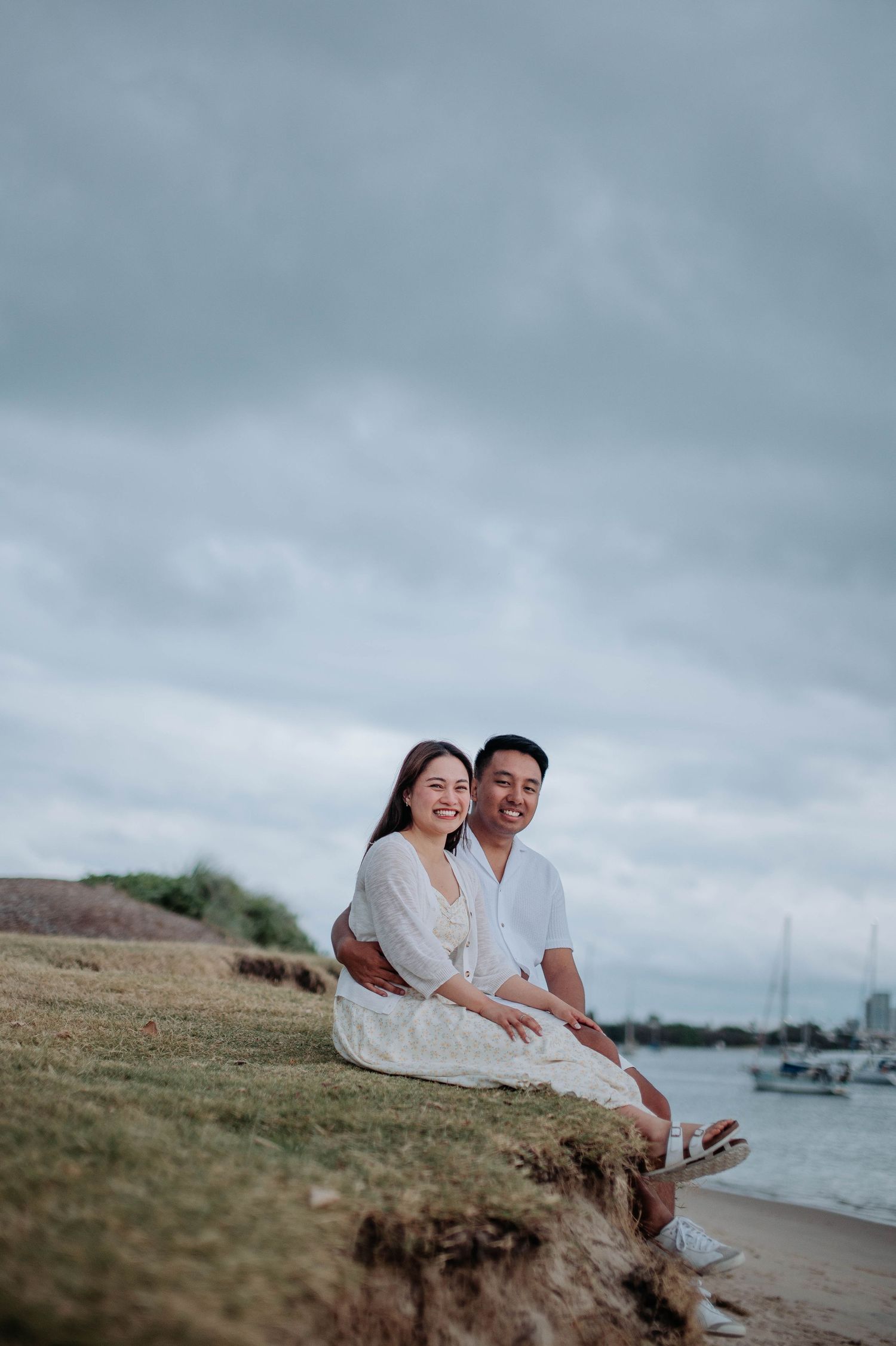 A couple shares a joyful moment while sitting on a grassy cliff by the ocean.