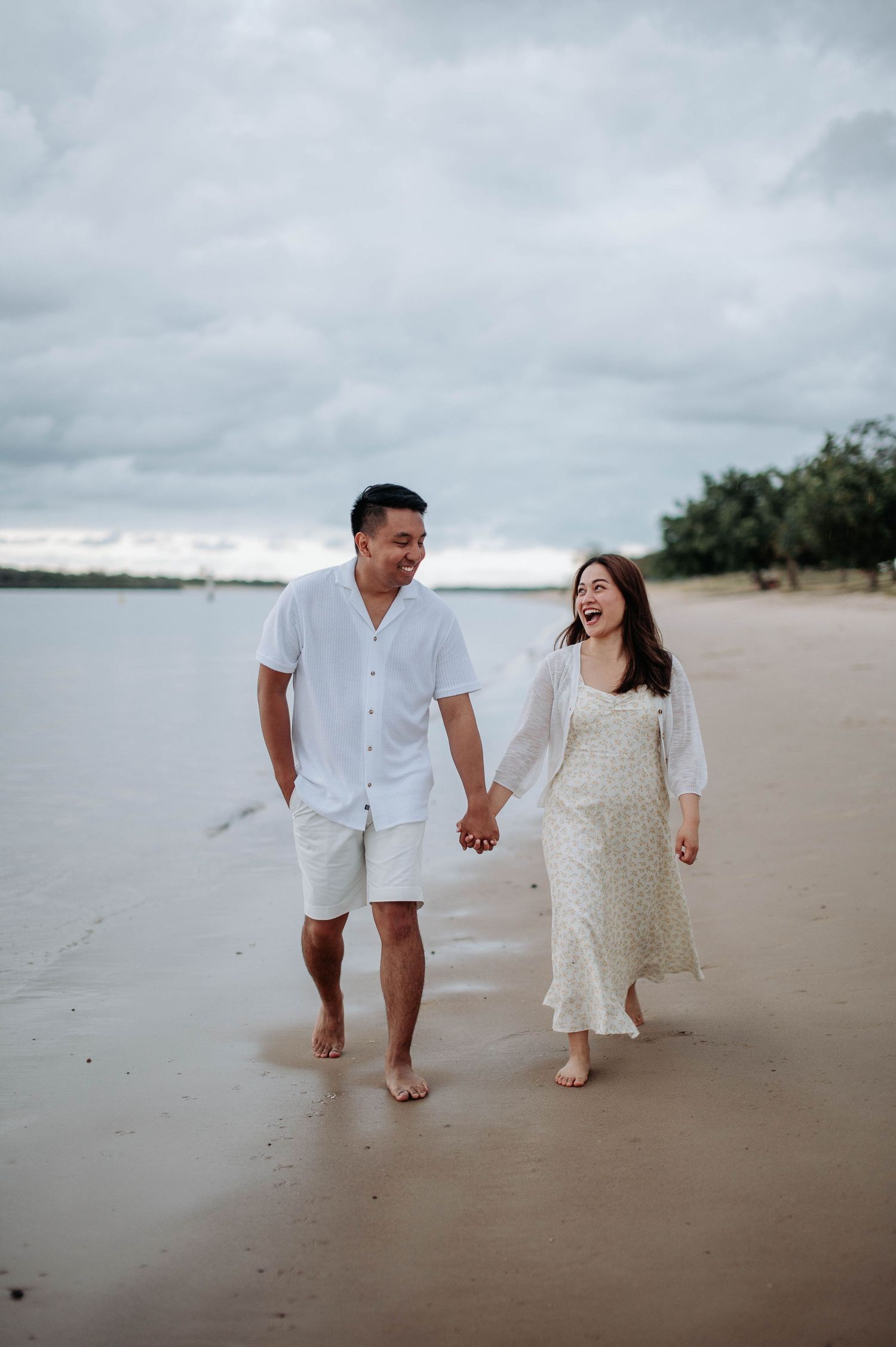 A couple walks barefoot along the beach shore under cloudy skies.