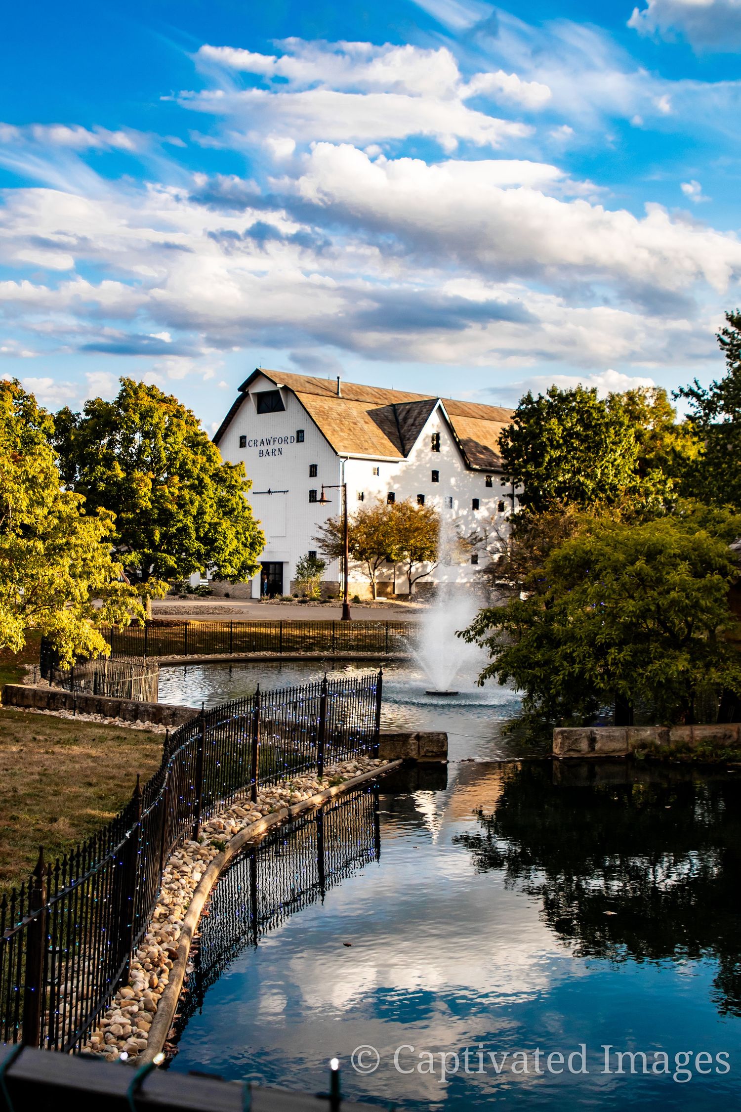 Historic white mill building with fountain in foreground reflecting in tranquil pond on sunny autumn day.