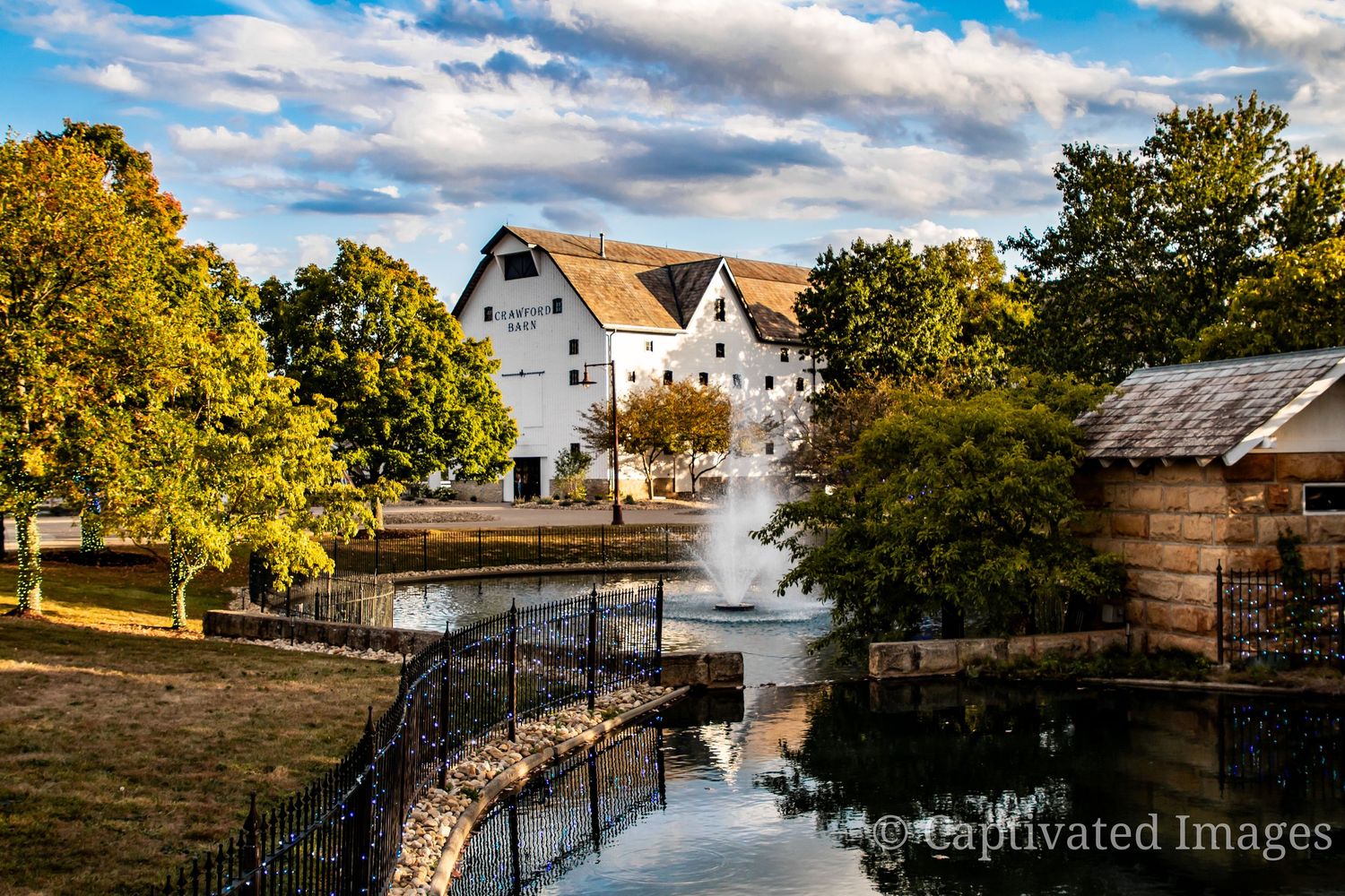 Historic white mill building reflects in calm water with curved stone bridge and autumn trees along riverbank.