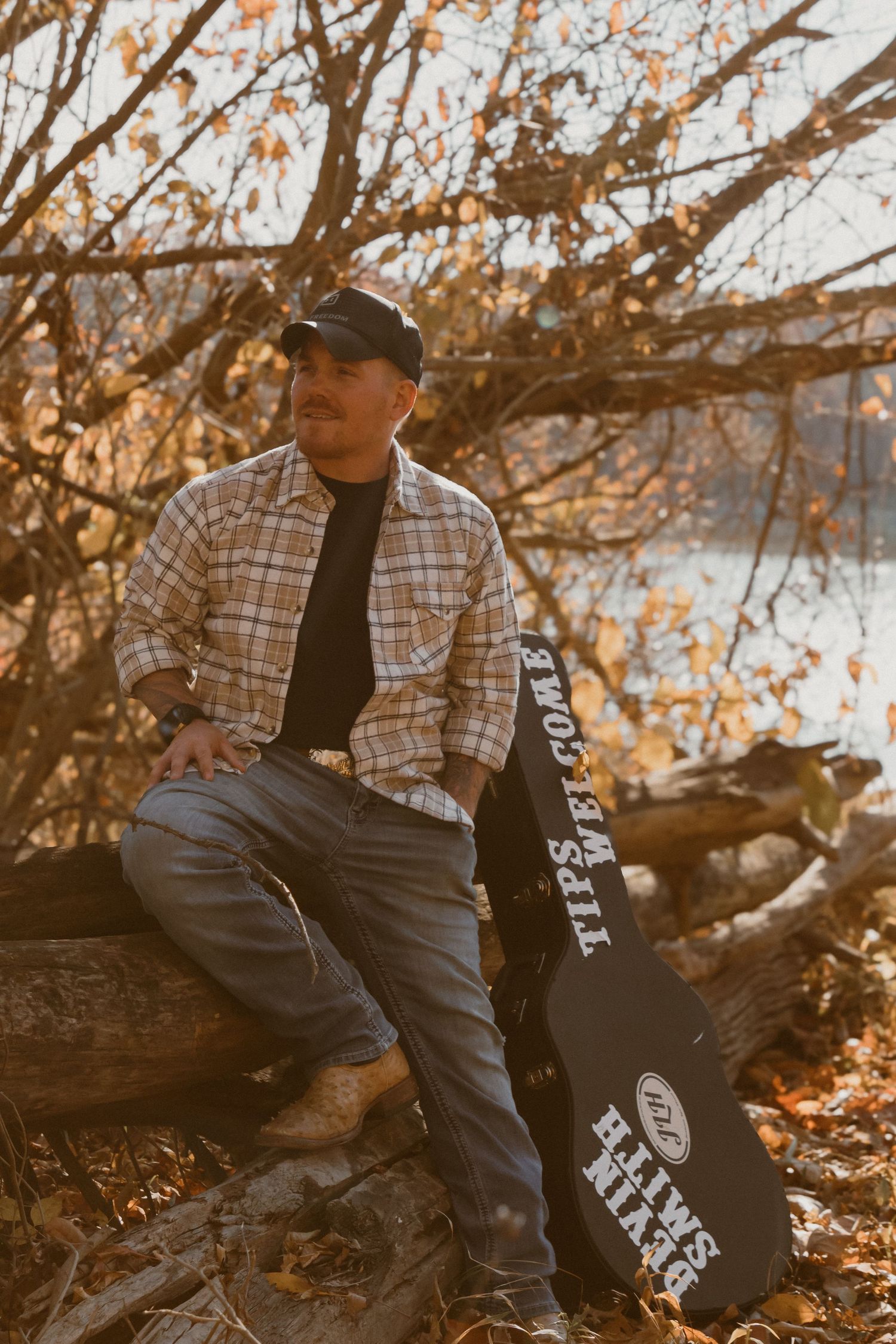 Musician sitting on fallen log with guitar case in autumn forest setting.