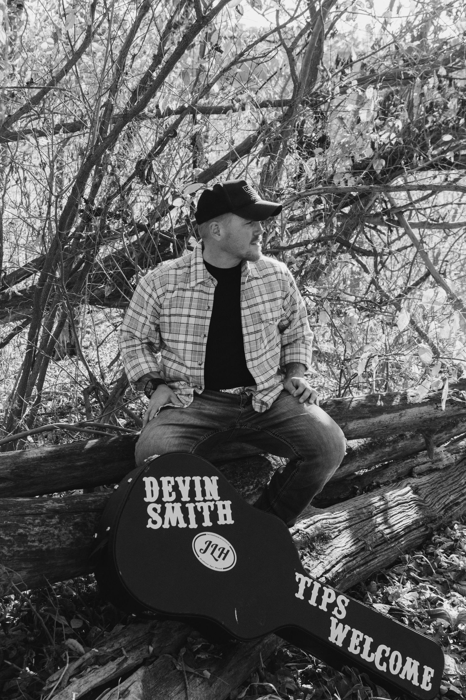 Black and white portrait of musician with guitar case in wooded setting.