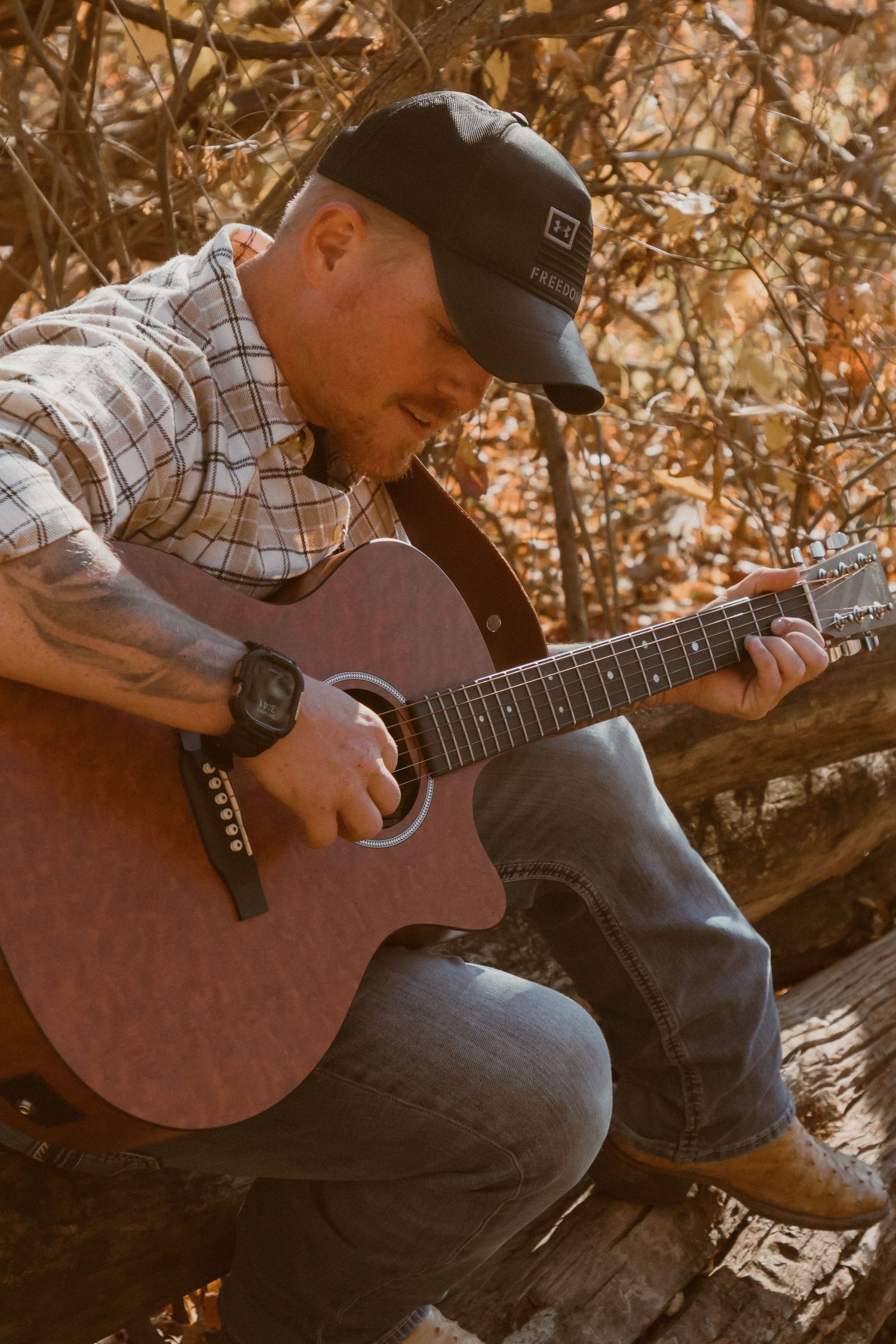 Guitarist playing acoustic guitar while sitting on fallen tree in warm autumn light.
