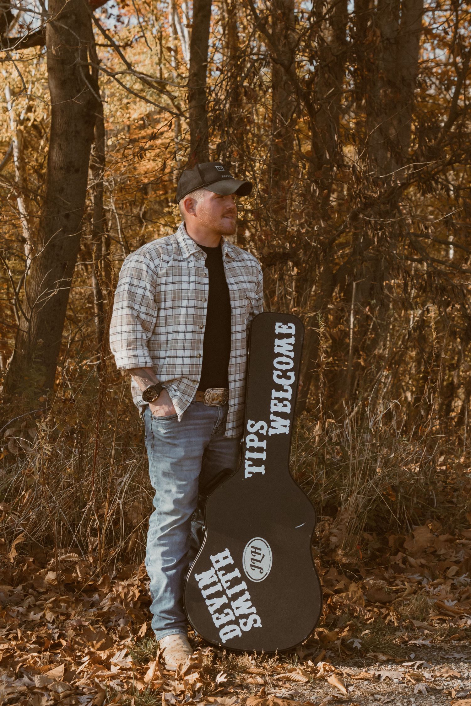 Country musician standing with guitar case against autumn forest backdrop.