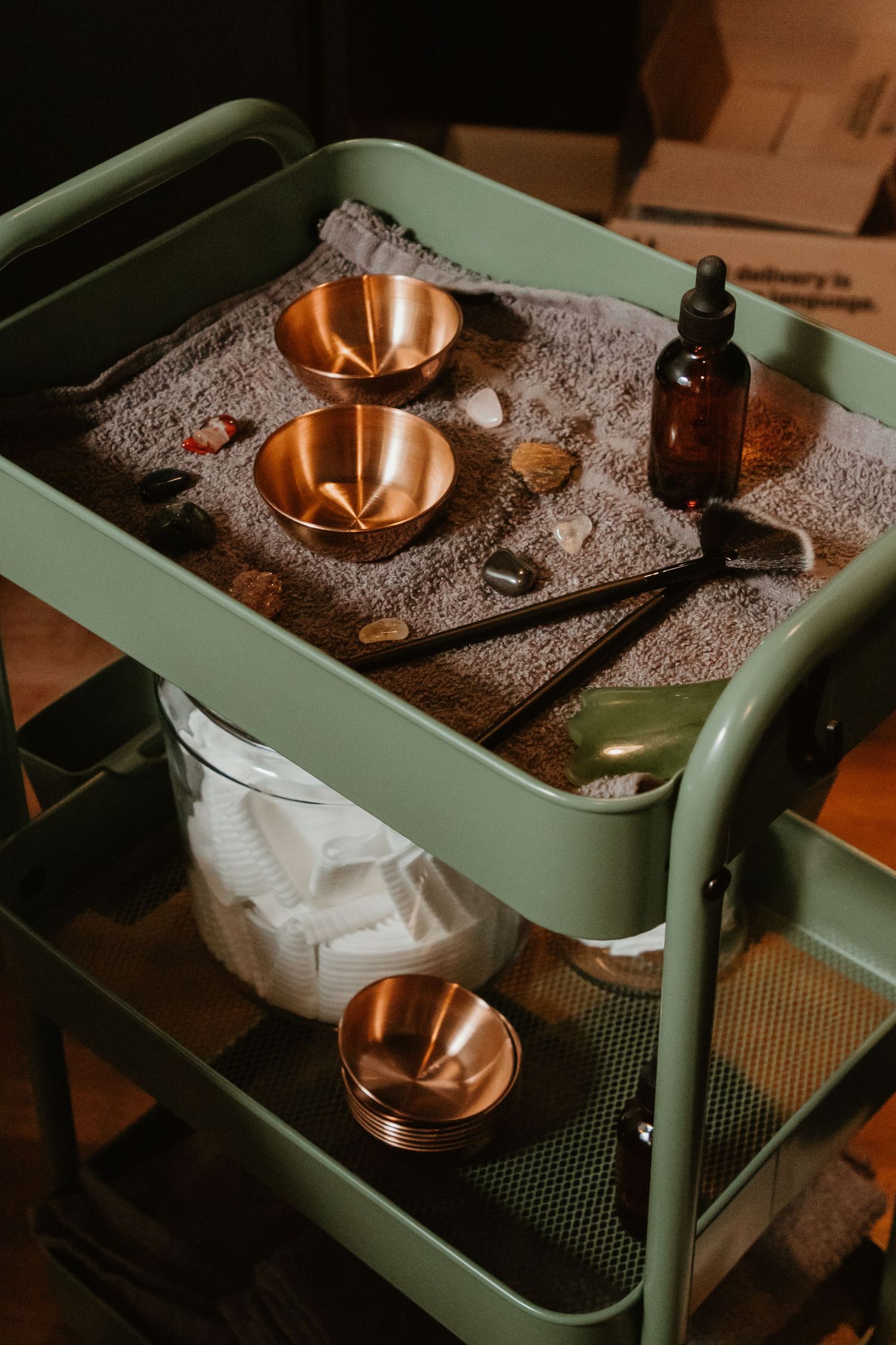 Vintage copper bowls and amber glass bottle arranged on sage green metal cart with textured gray surface.