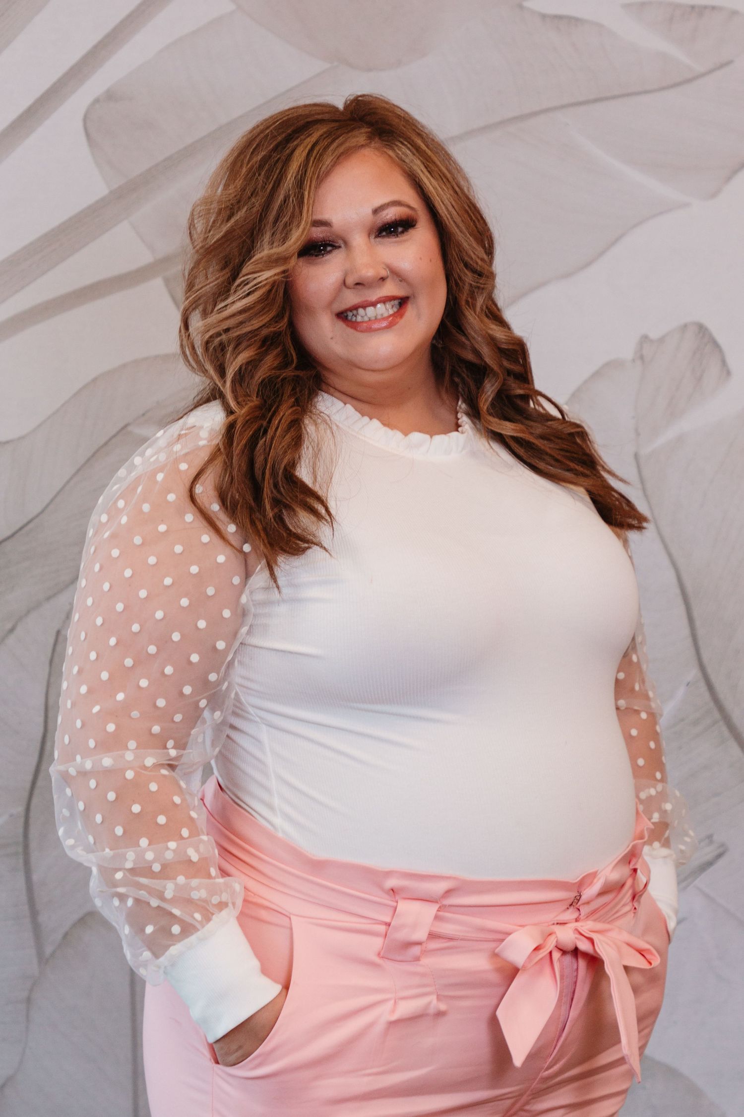 Portrait of a person in a white top with sheer polka dot sleeves and pink pants.