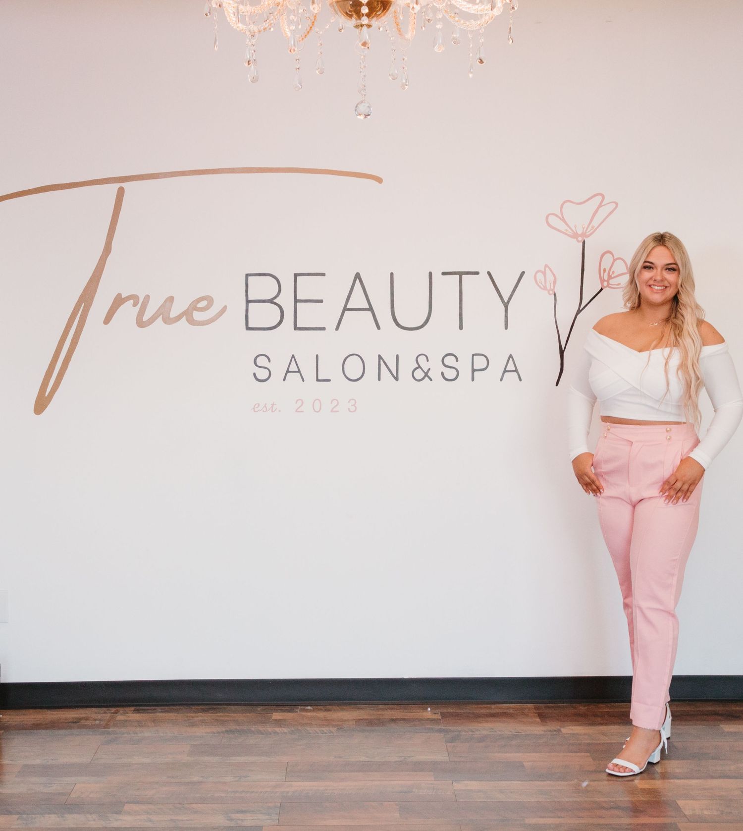 Professional standing by True Beauty Salon & Spa wall signage wearing pink and white attire.