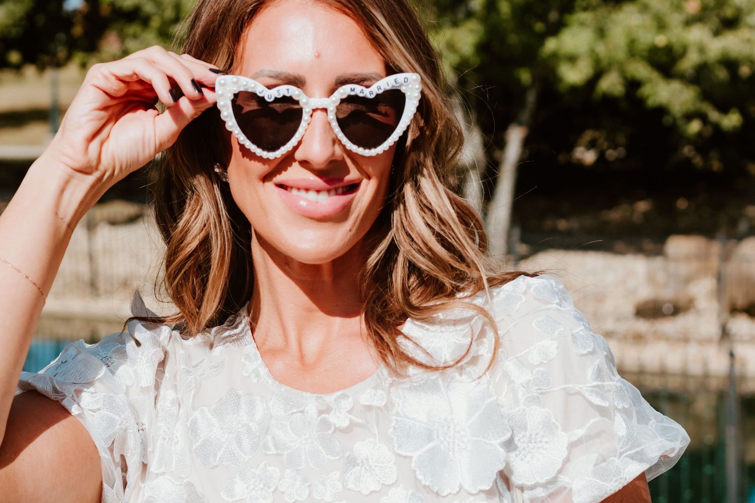 Fun bridal summer portrait wearing white lace dress and heart-shaped sunglasses in sunny outdoor setting.