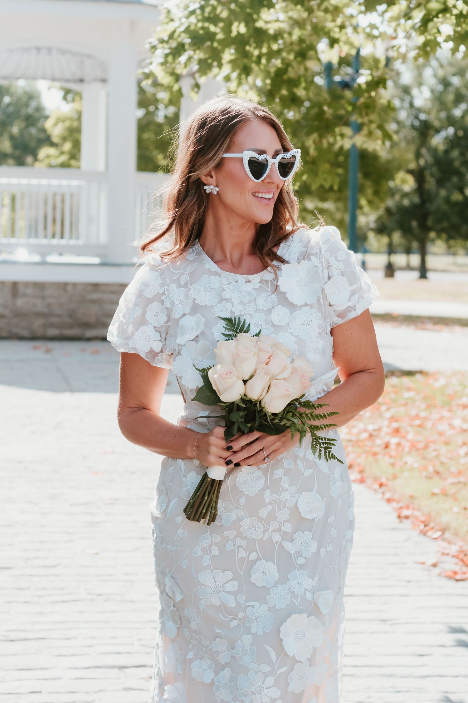 Elegant bride in white floral dress holding bouquet of white roses near gazebo.