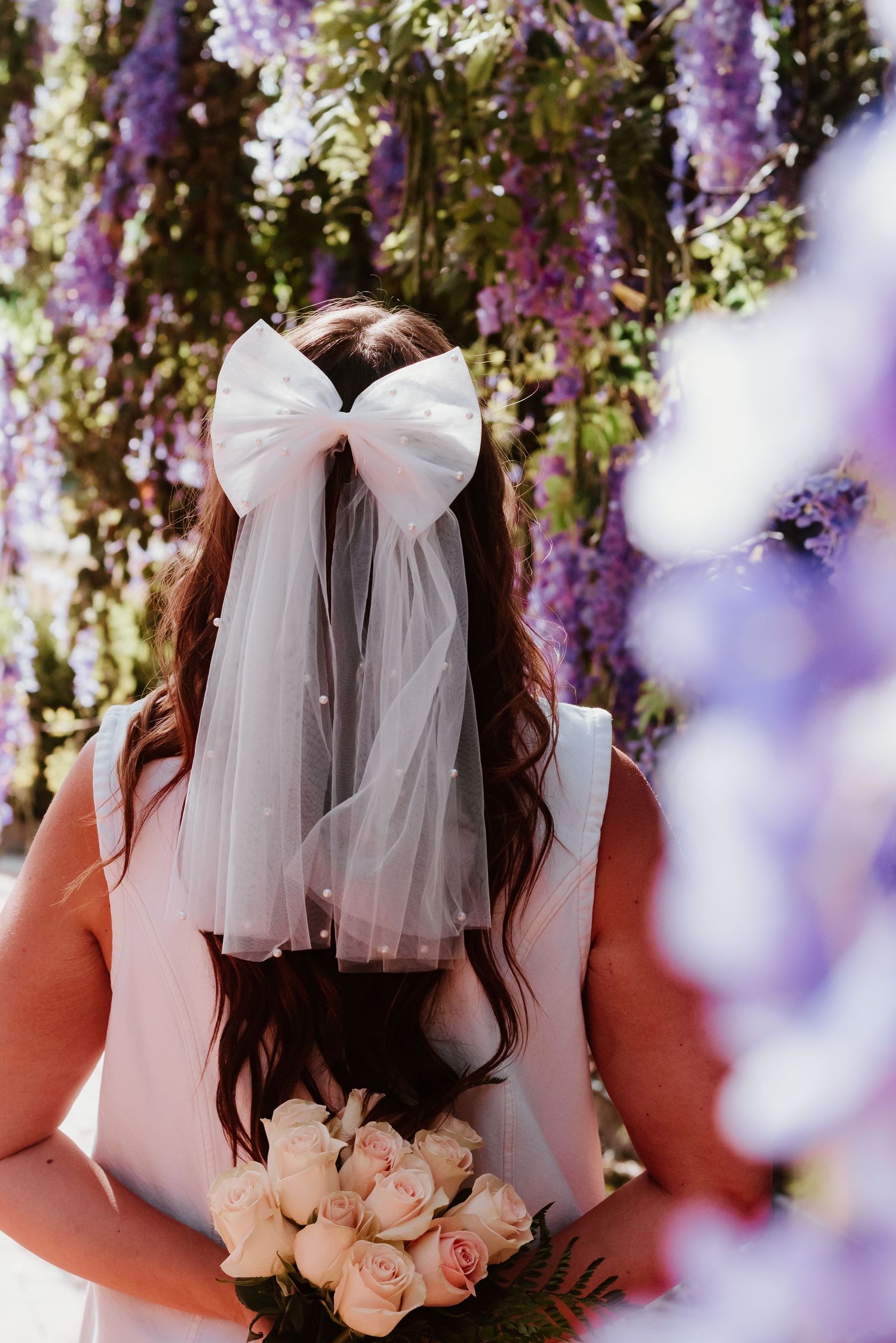 Back view of white bow veil and roses against purple wisteria flowers.