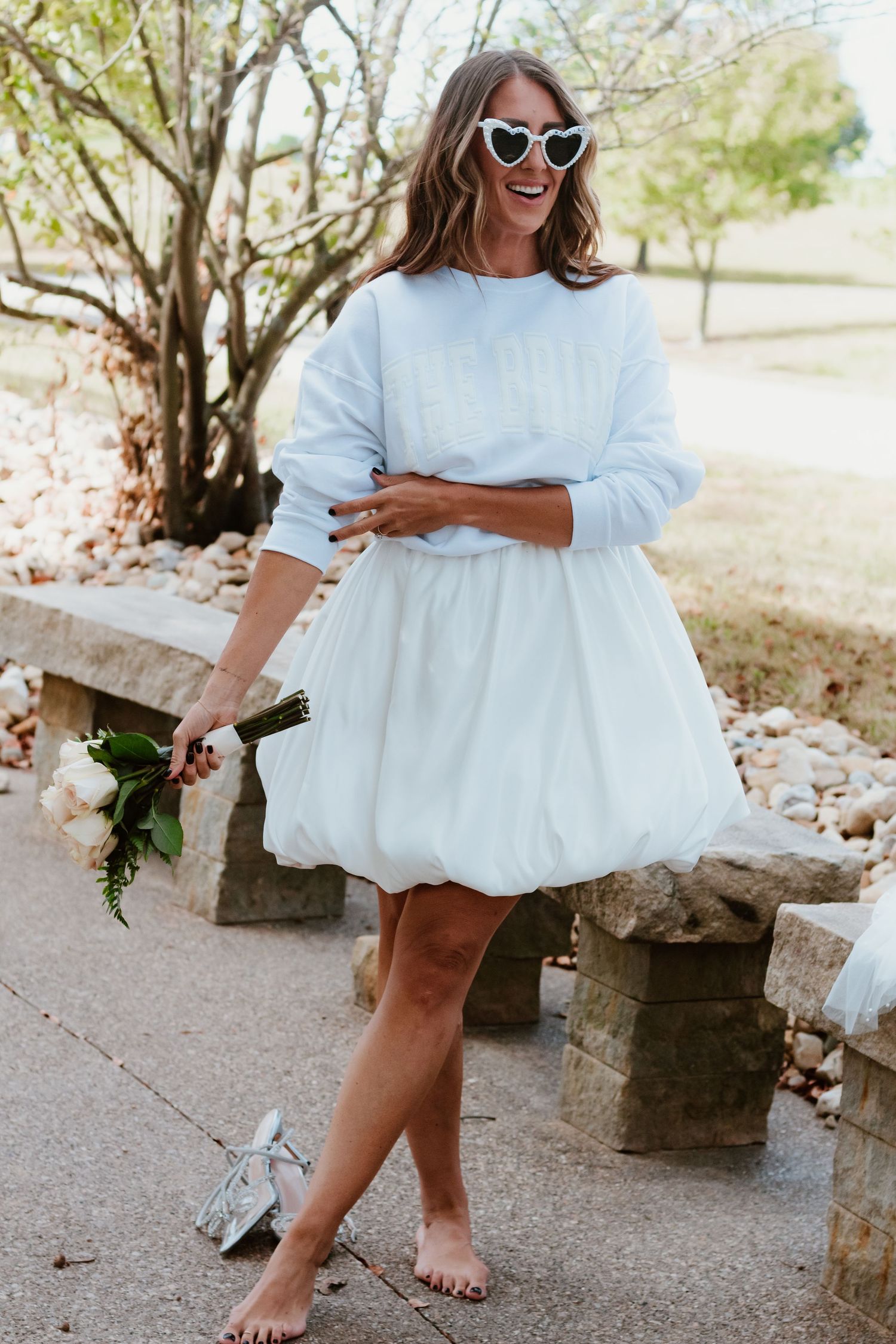 Barefoot pose in white sweater dress holding floral bouquet on stone wall.