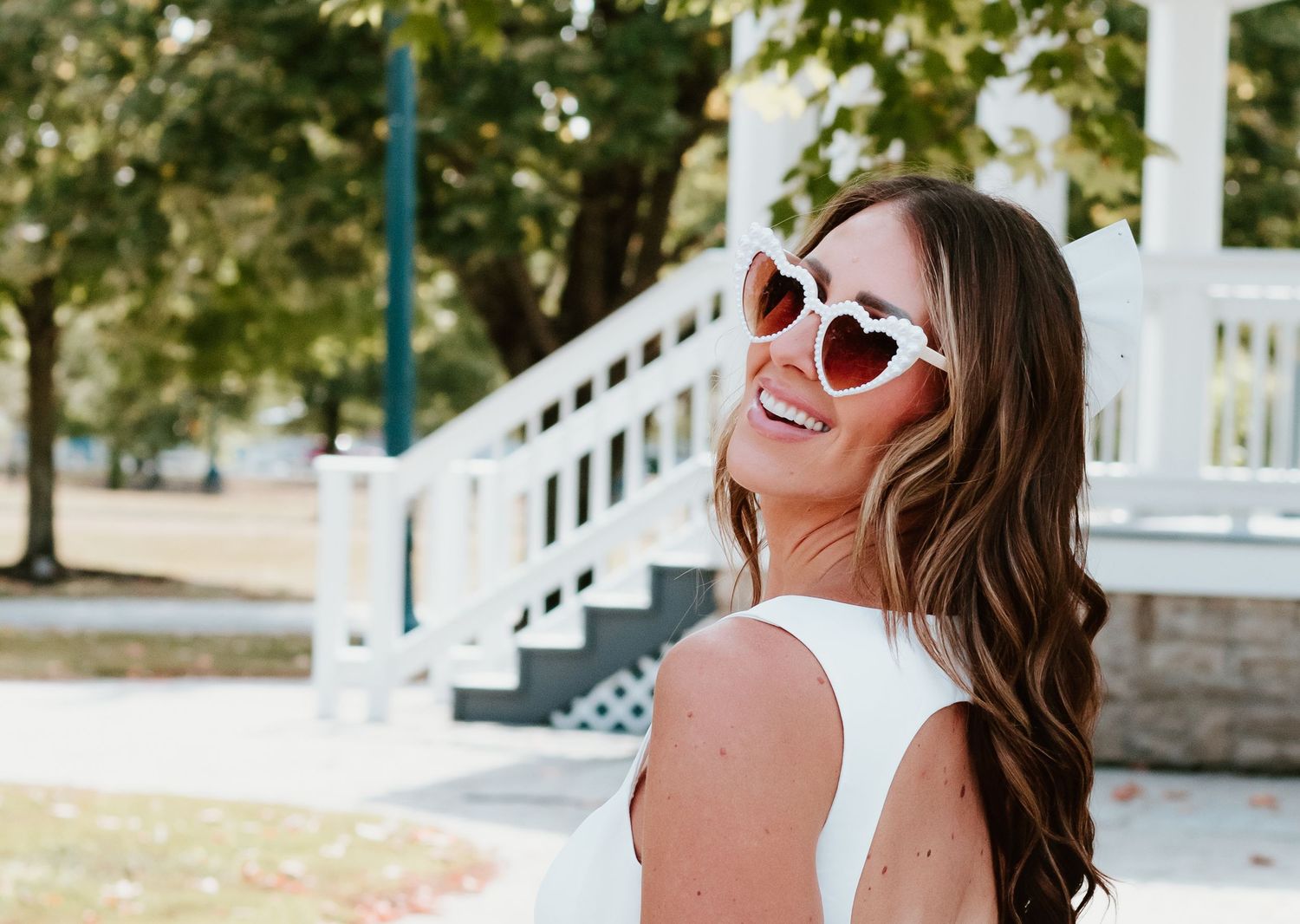 Joyful summer portrait wearing retro white sunglasses and white dress against outdoor backdrop.