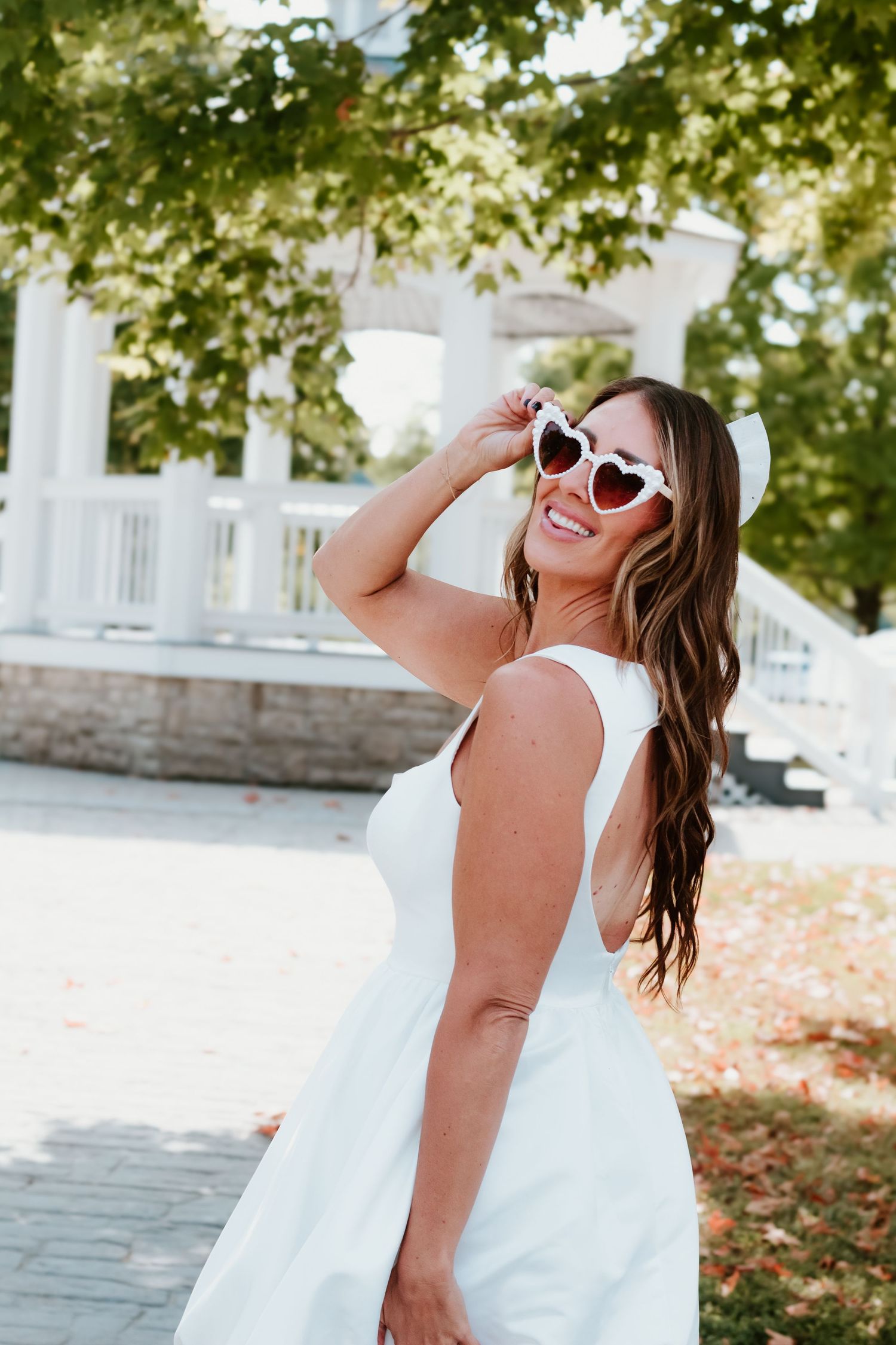 Summer bride in white dress and heart-shaped sunglasses posing by gazebo.
