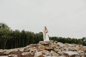 A person in a white dress poses elegantly on top of a rustic stone wall against a lush forest backdrop.