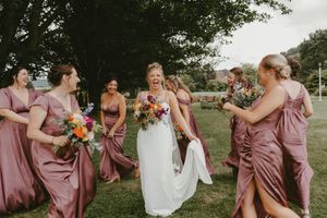 Wedding party wearing mauve bridesmaid dresses gather outdoors on a lawn with colorful bouquets.