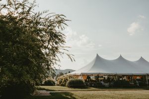 White outdoor wedding tent set up at dusk in a grassy field with trees and mountains in the background.