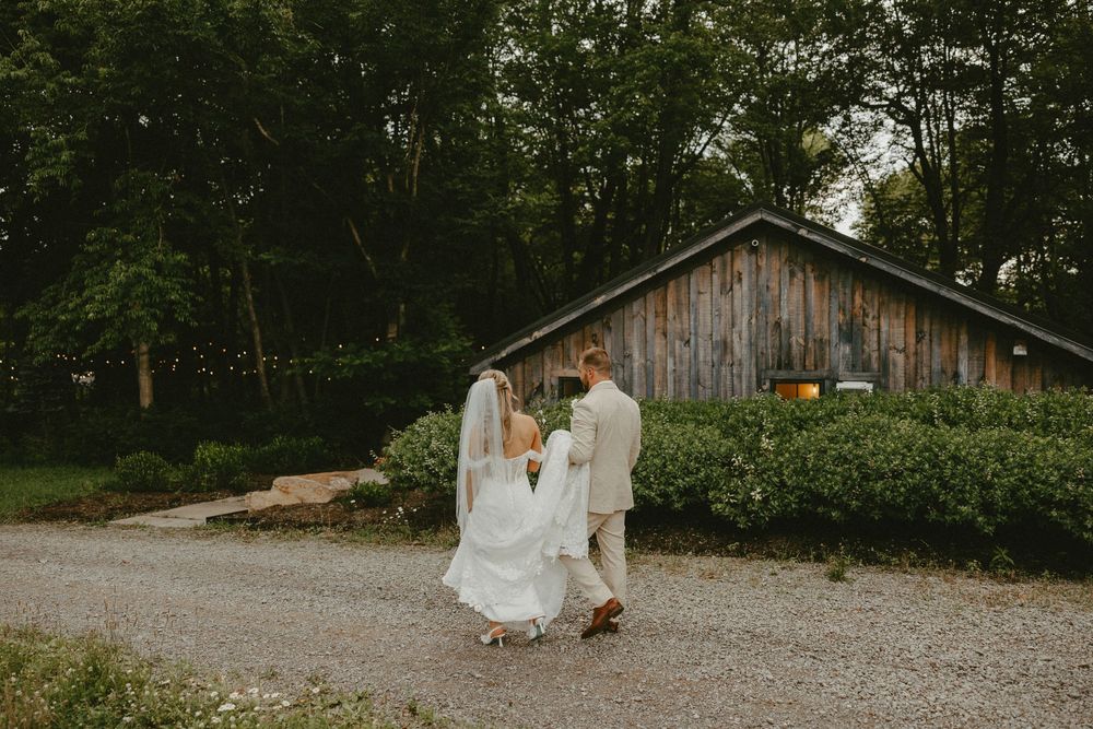 A bride and groom share a romantic moment outdoors near rustic wooden barn structures surrounded by lush green trees.
