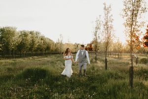 A couple walks together through a sun-lit meadow during golden hour with tall grass and trees in the background.