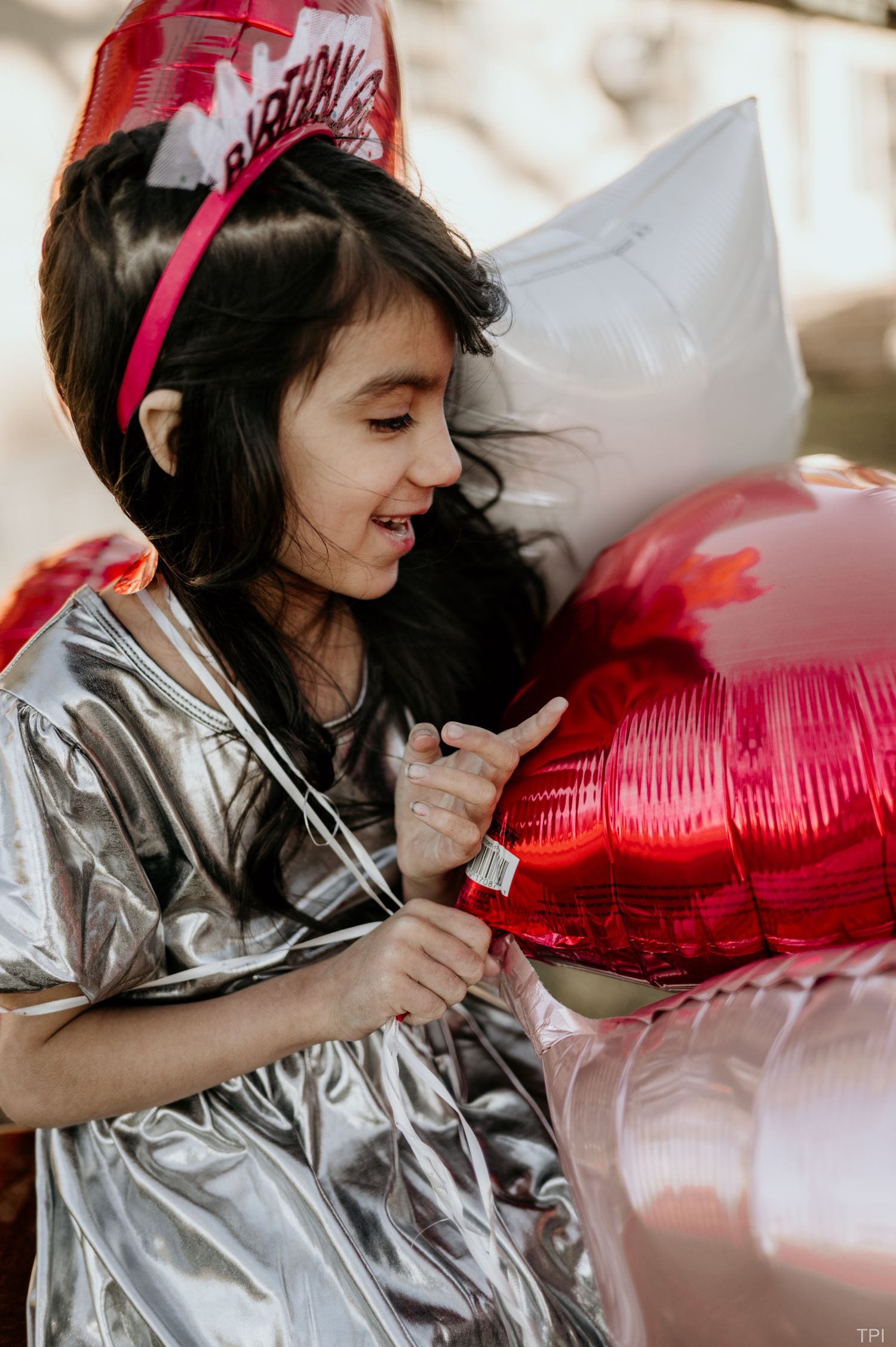 Person wearing silver outfit and crown poses with heart-shaped red balloons in celebratory setting.
