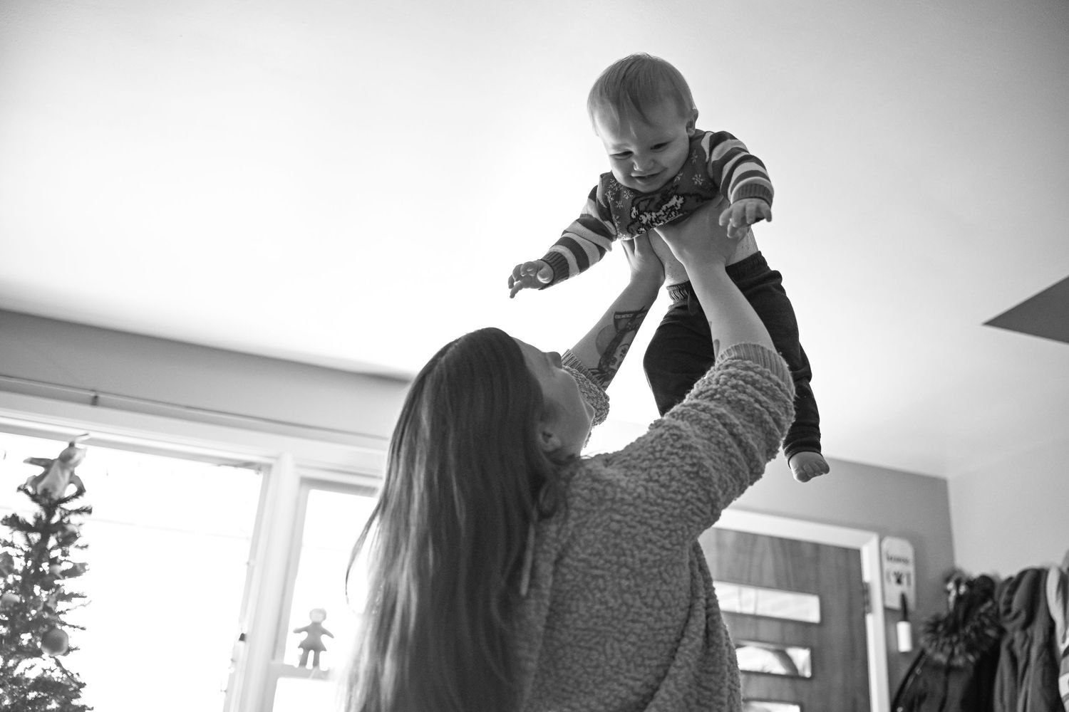 A black and white photo of a woman standing in her living room, lifting her baby boy into the air above her head to make him laugh