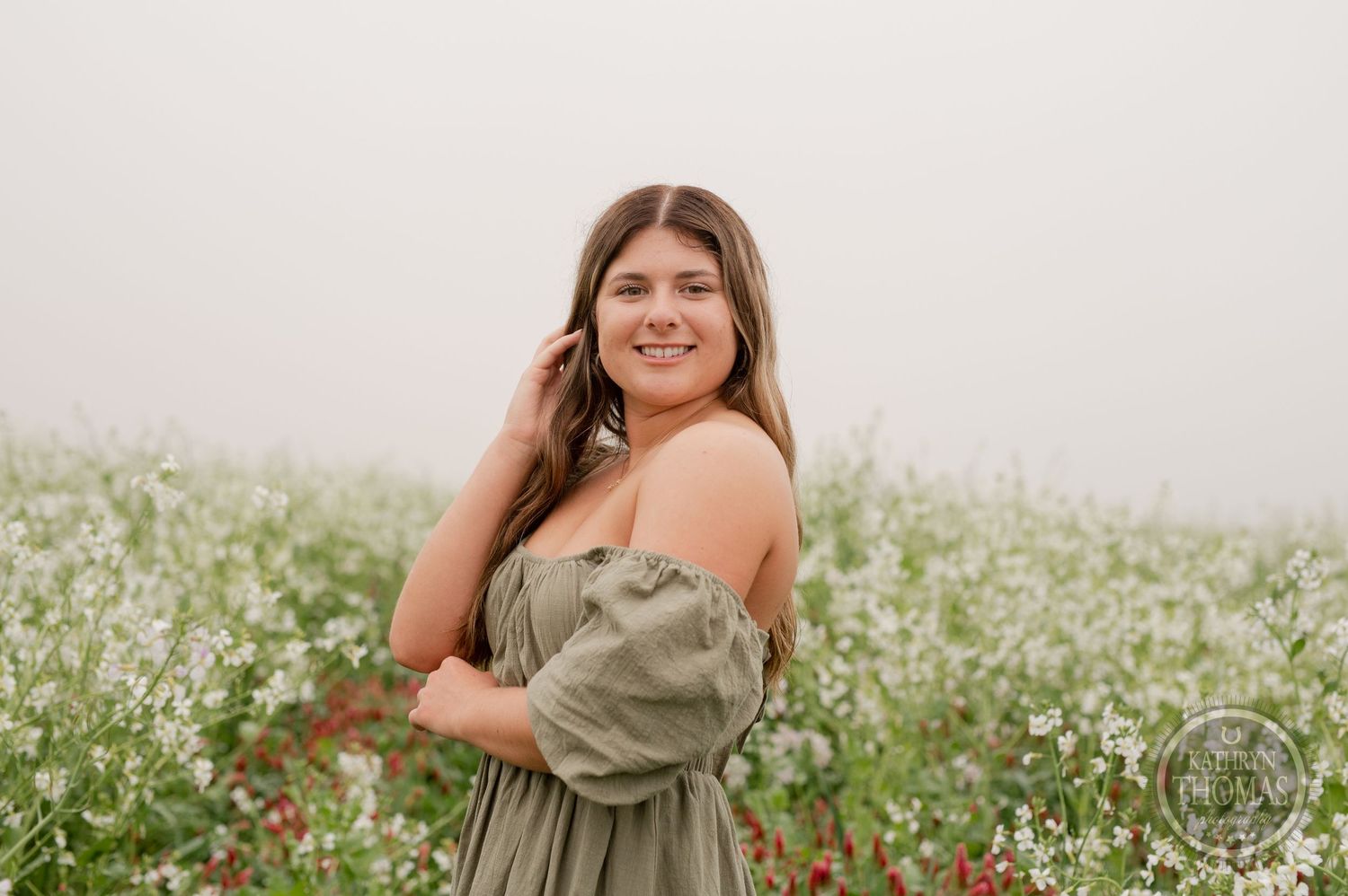 Person in a green dress smiles while standing among blooming white wildflowers in a misty field.
