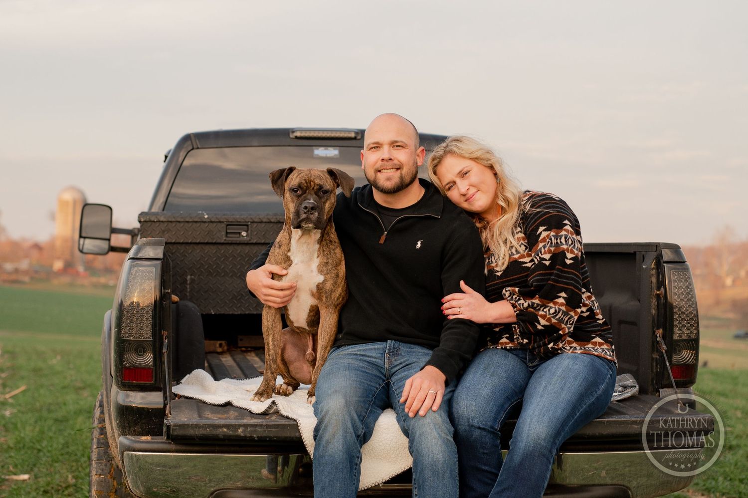 A couple and their two dogs pose together in the bed of a black pickup truck at sunset.
