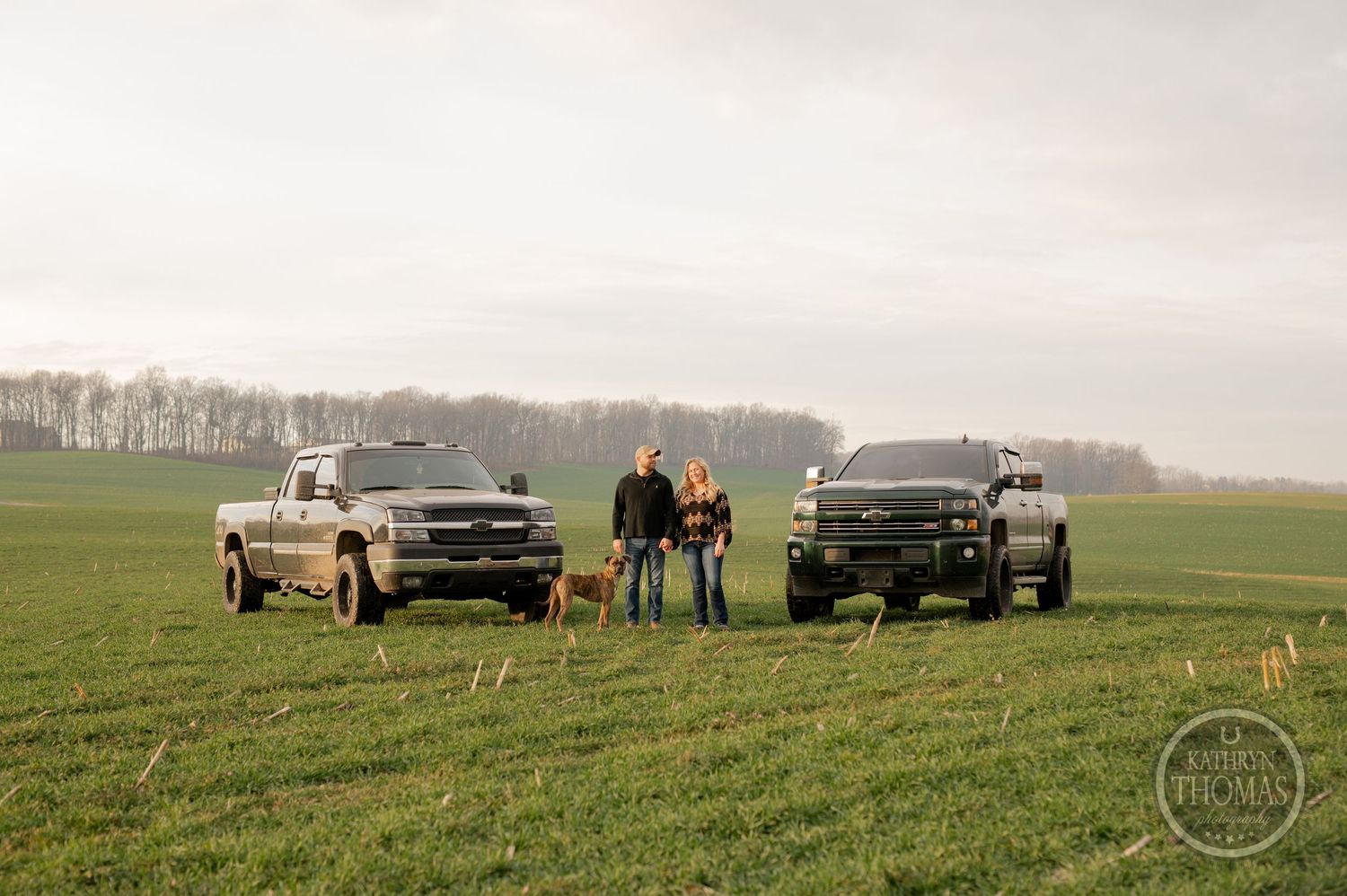 Two couples stand between parked pickup trucks in a wide-open green field.