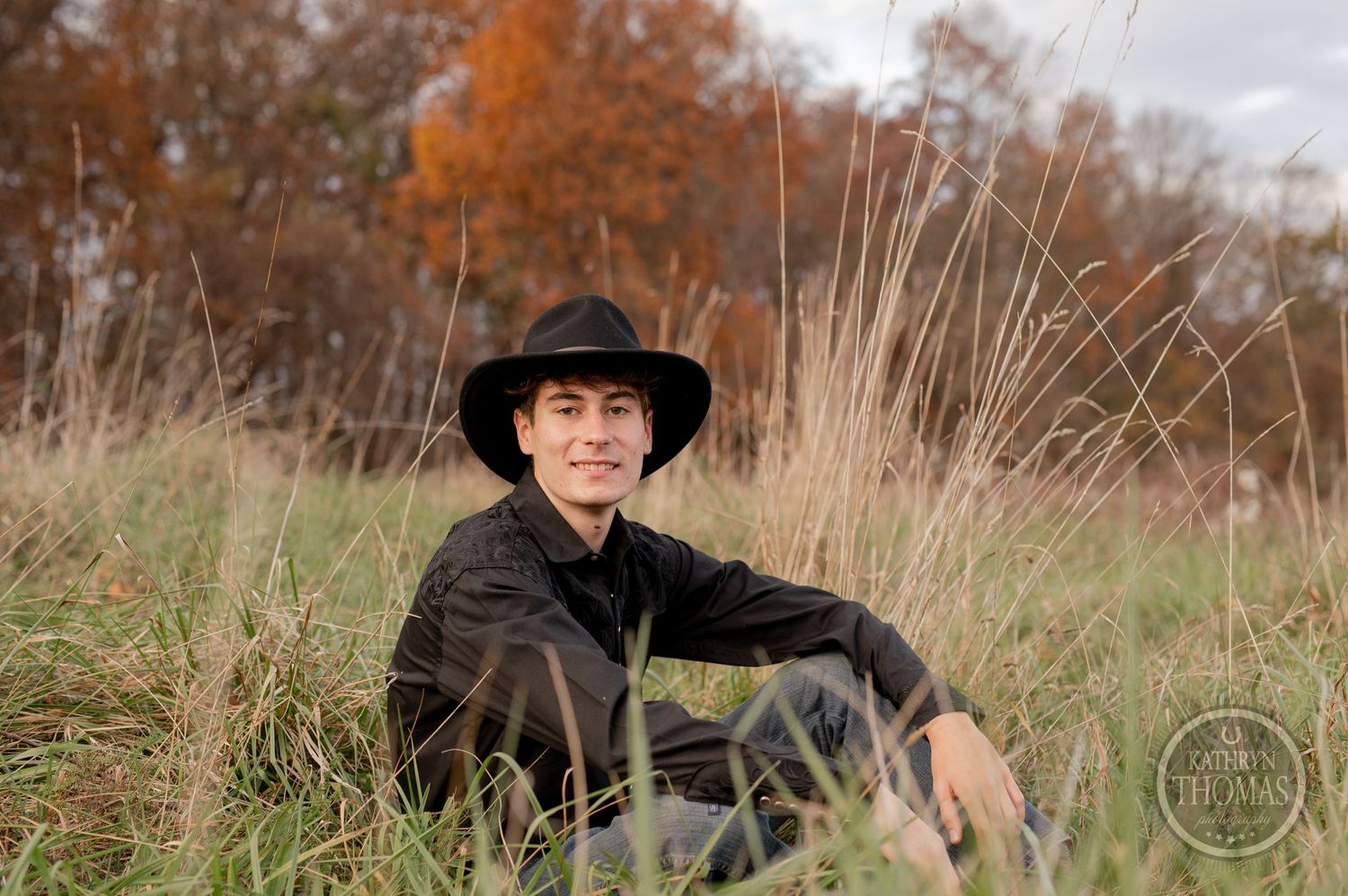 Casual western-style pose in tall grass field with autumn colors in the background.