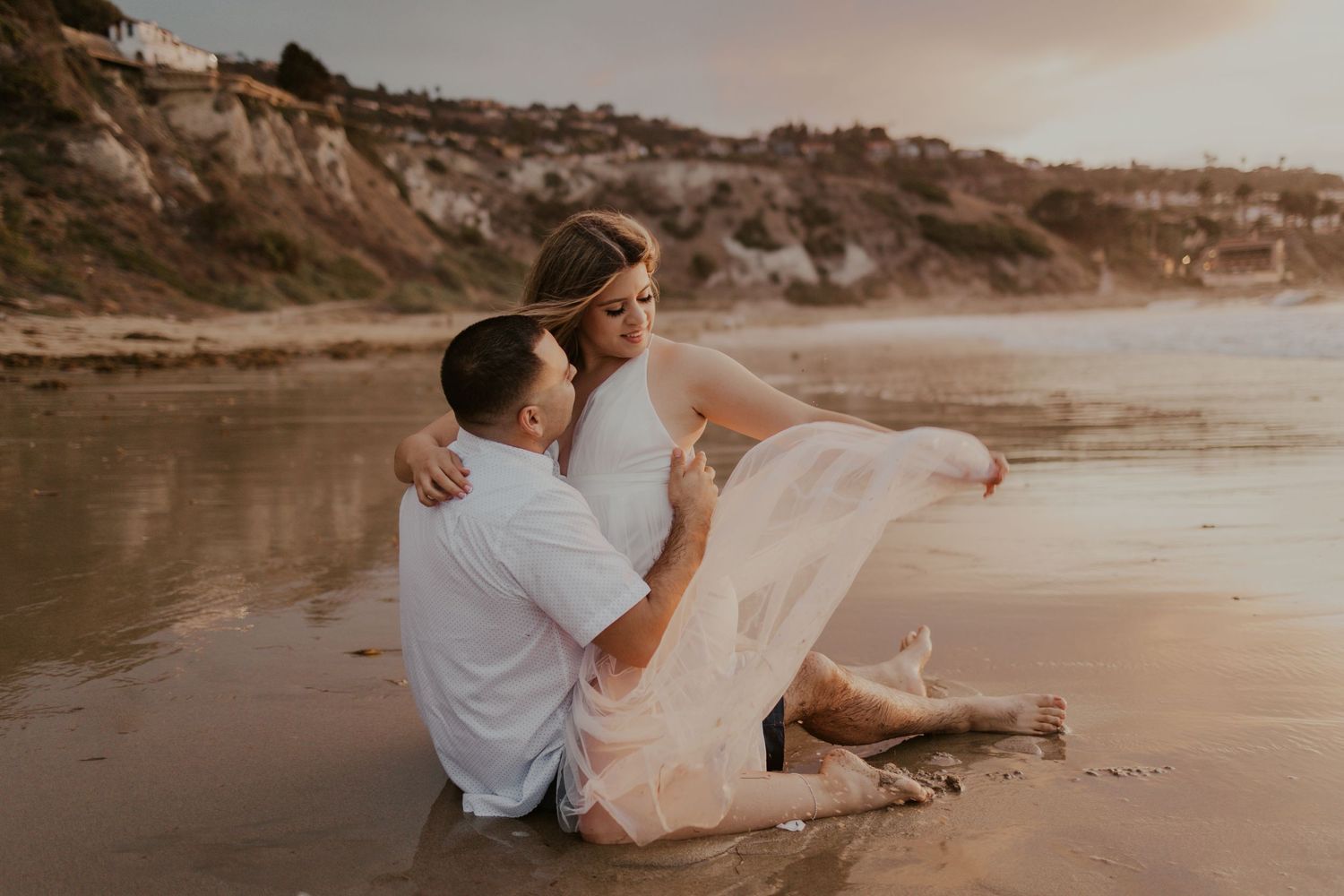 Romantic sunset beach photoshoot with couple sitting on sand as dress flows in the breeze.