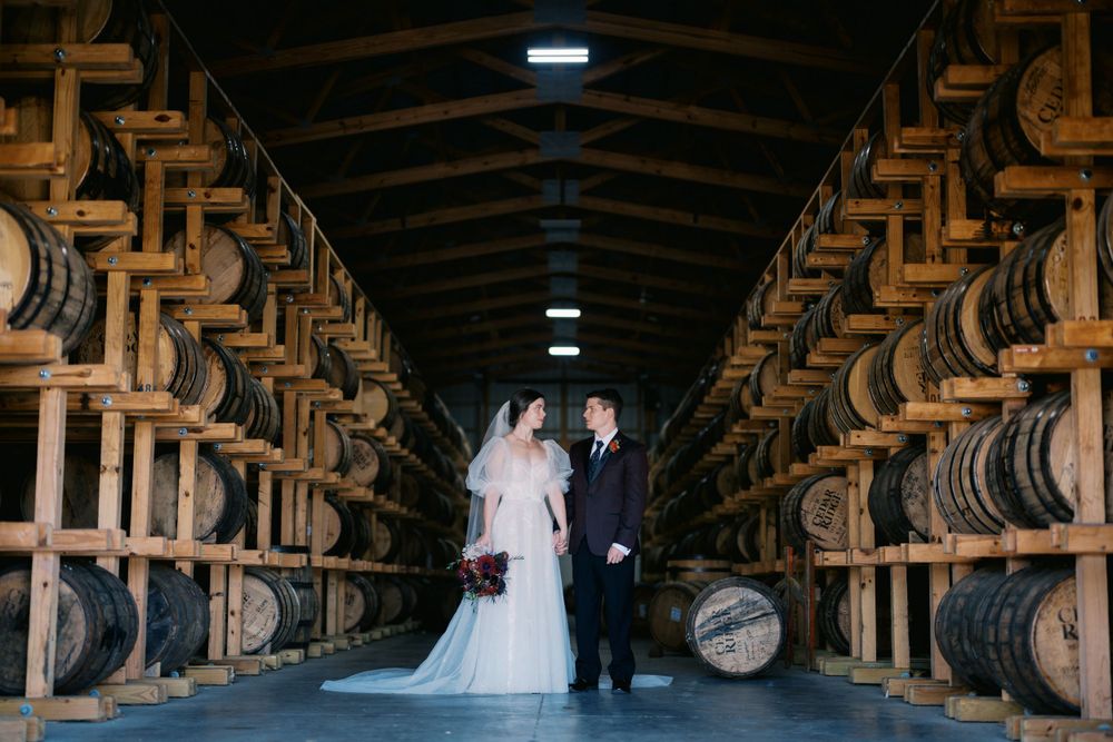 A couple poses amid rows of wooden whiskey barrels in a dark rustic bourbon distillery warehouse.