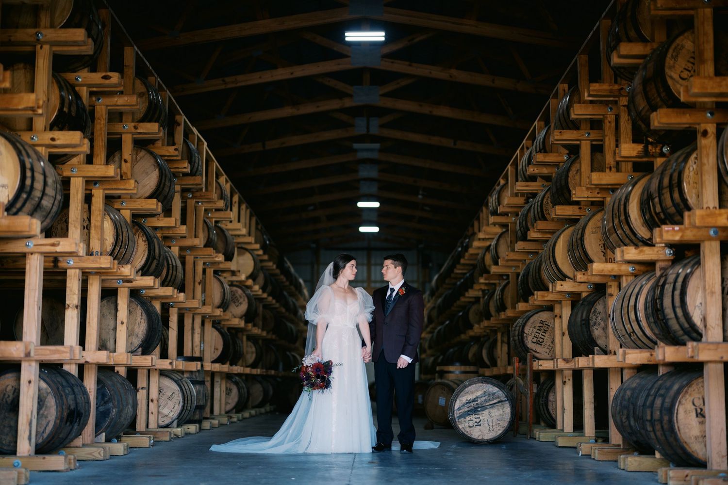 A couple poses amid rows of wooden whiskey barrels in a dark rustic bourbon distillery warehouse.