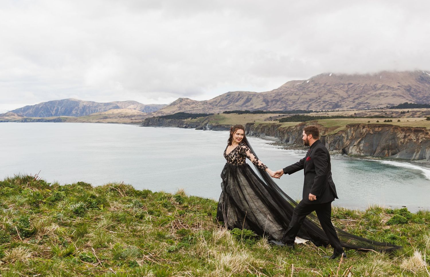 Two people adjust a flowing black dress against a dramatic coastal cliff backdrop.
