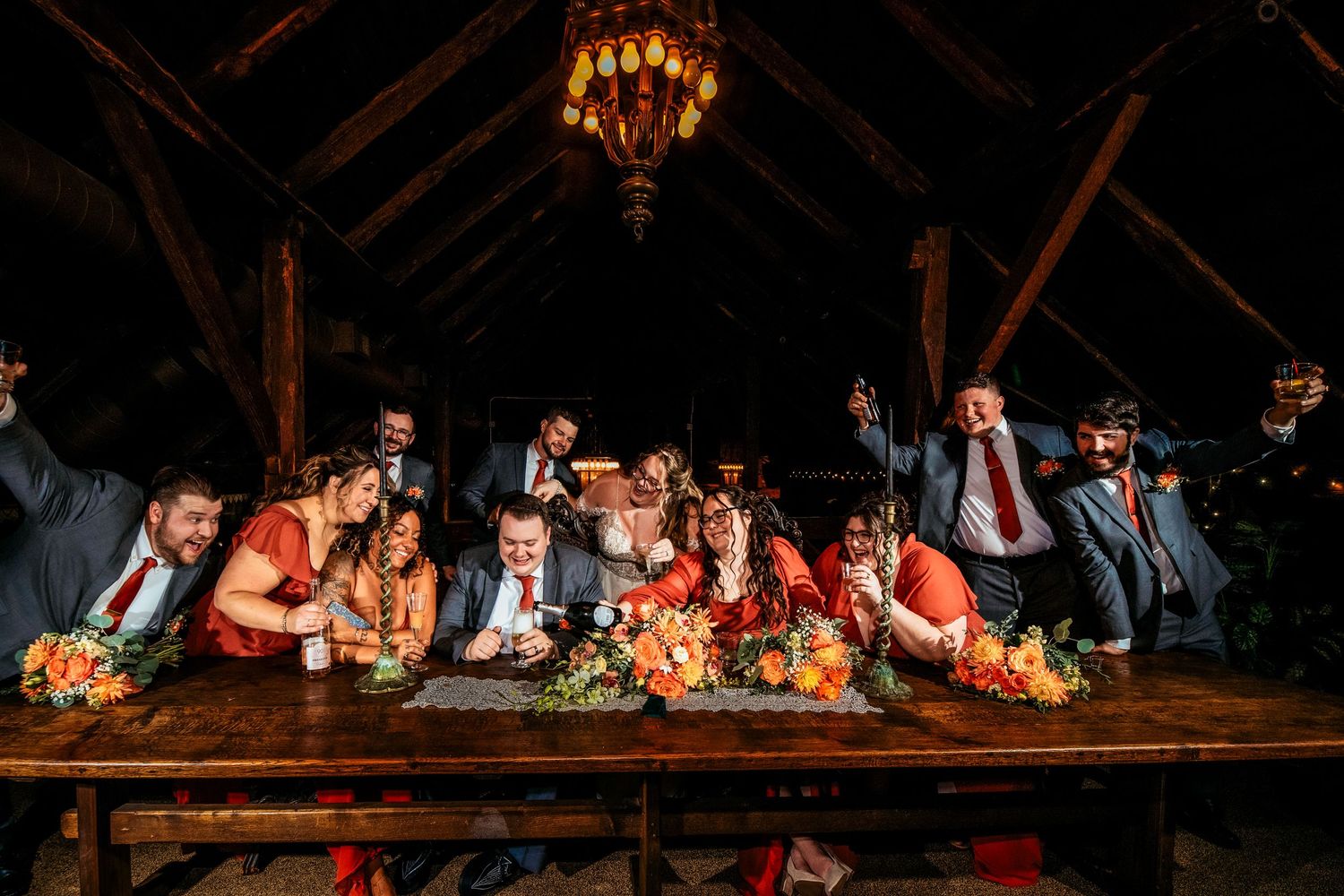 Wedding party gathered around a rustic wooden table under a chandelier in a dimly lit barn venue.