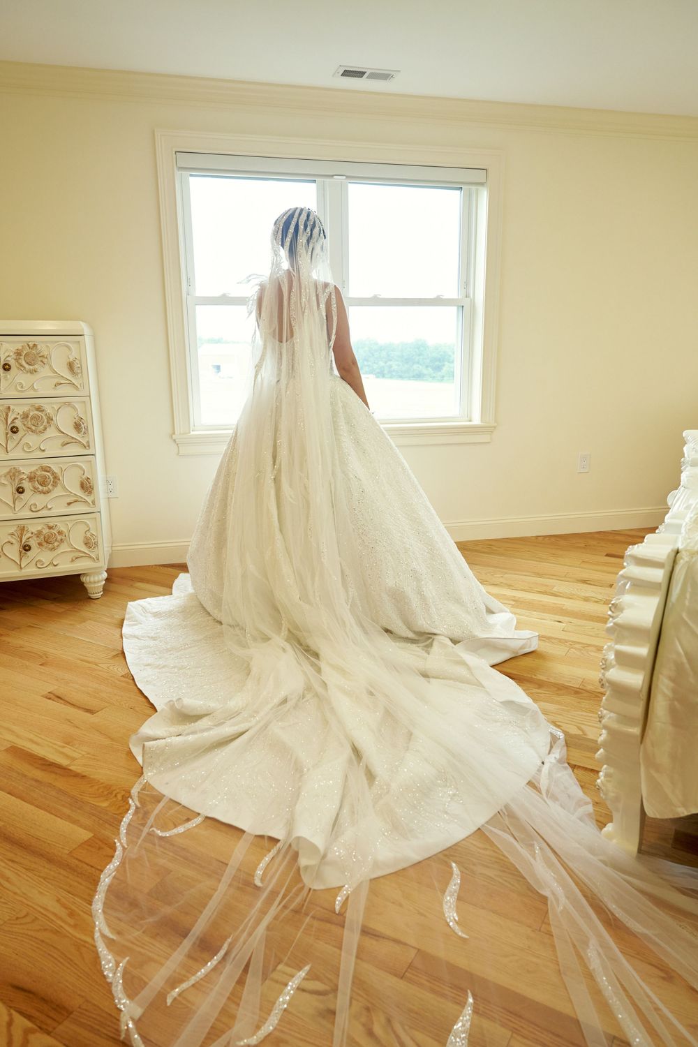 Flowing white wedding gown with train displayed on hardwood floor in bright room.