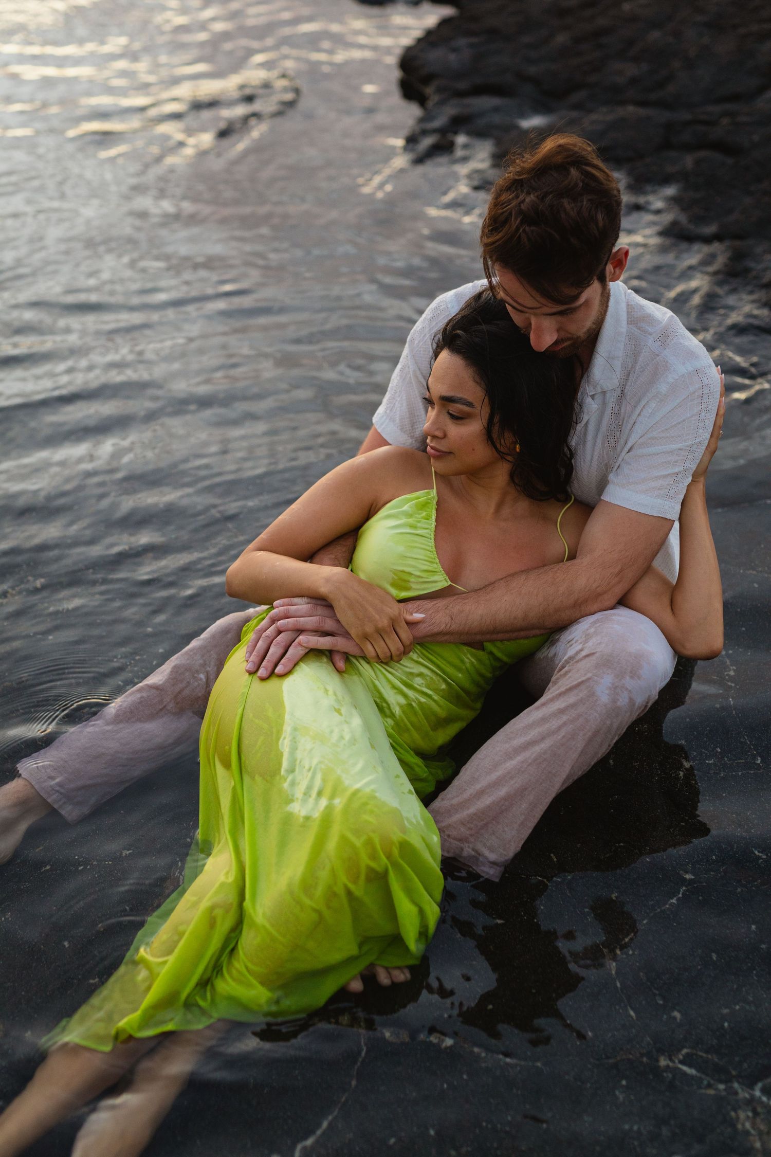 Couple sitting together in shallow ocean water at sunset, woman in flowing green dress leaning against partner.