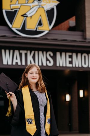 A graduate in cap and gown with a gold honors stole stands smiling outside Watkins Memorial building.