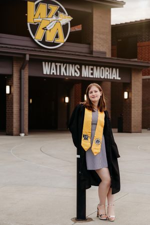 A graduate in cap and gown poses outside Watkins Memorial building entrance with school logo visible above.