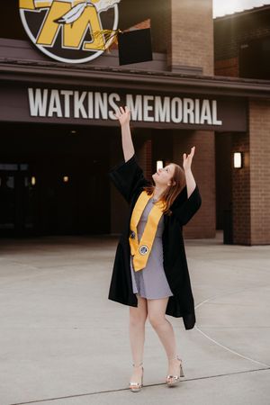 Graduate wearing black gown and gold heels poses in front of Watkins Memorial sign for graduation photos.