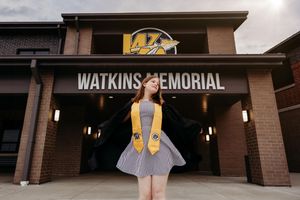 Graduate stands in front of Watkins Memorial building entrance wearing a gray dress and yellow stole.