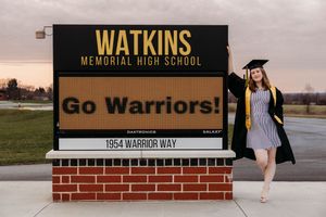 Graduate in cap and gown poses by Watkins Memorial High School Warriors sign at sunset.