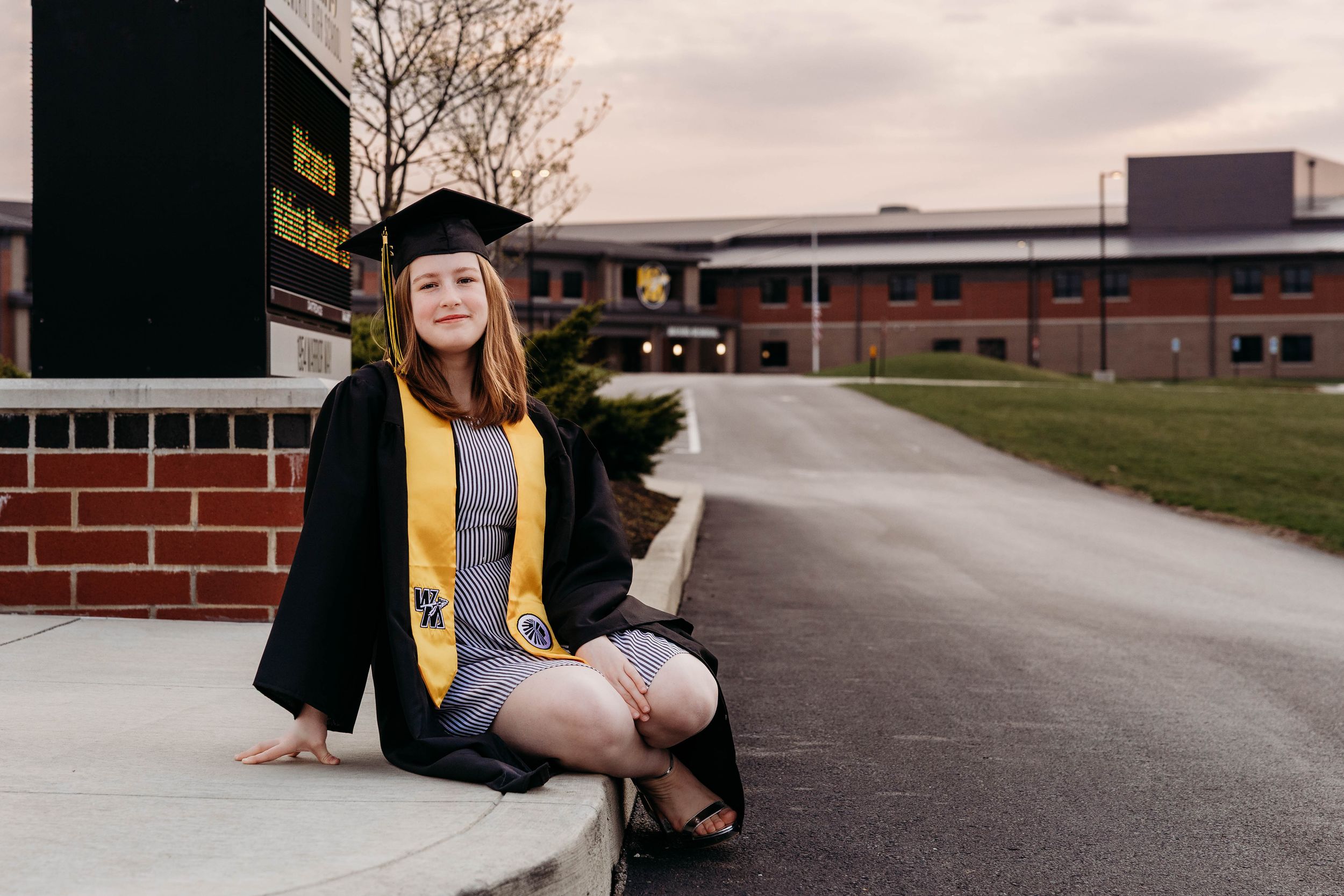 Graduate in academic regalia sits on brick wall outside brick school building.