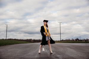 Graduate in black cap and gown with gold stole poses on empty road under cloudy sky.