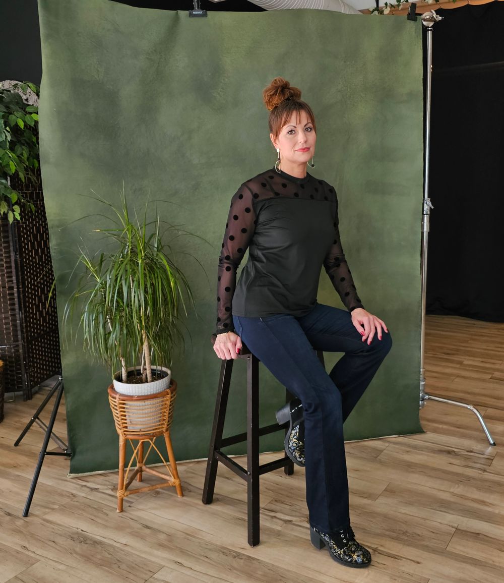 A figure sits on a tall bar stool against a green backdrop in a photo studio setting with hardwood floors and potted plants.