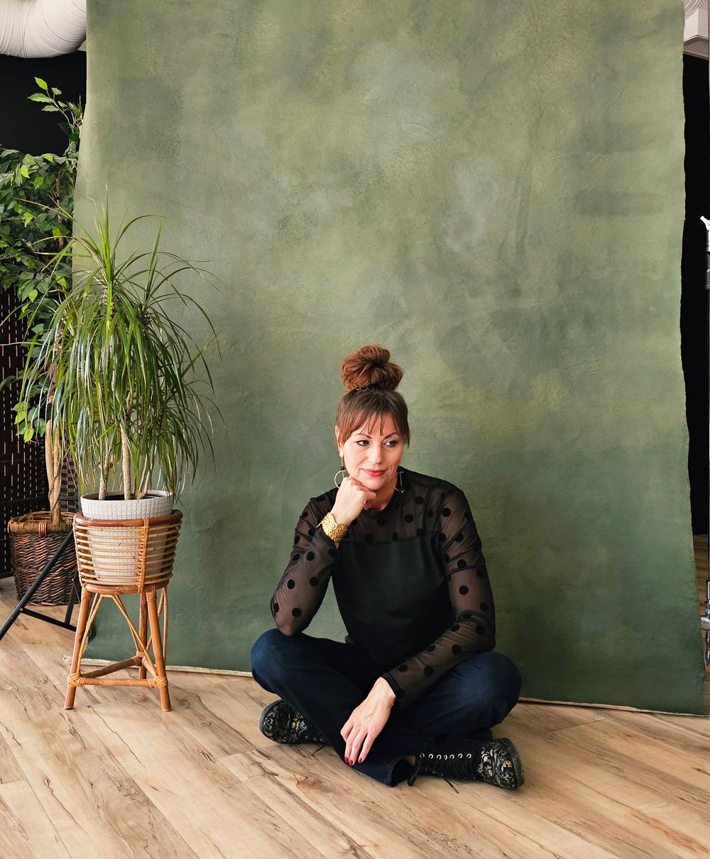 A figure in black attire lies on a wooden floor next to a potted plant against a green textured wall.